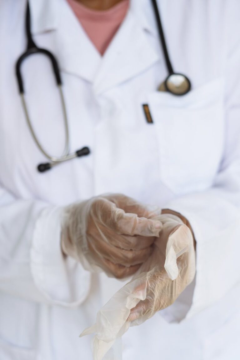 Faceless ethnic medical worker in lab coat and stethoscope taking of transparent gloves after approaching patients for examination in modern hospital