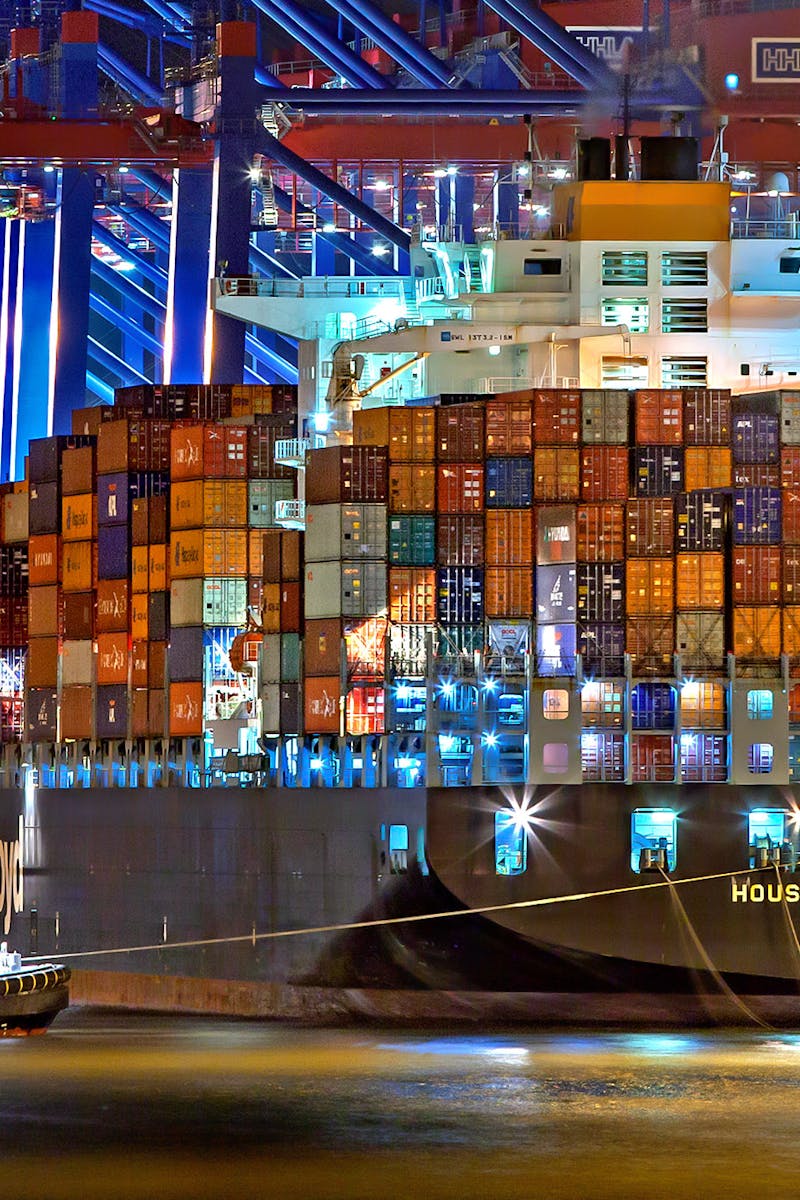 A brightly lit cargo ship at Hamburg harbor with stacked containers and a tugboat.